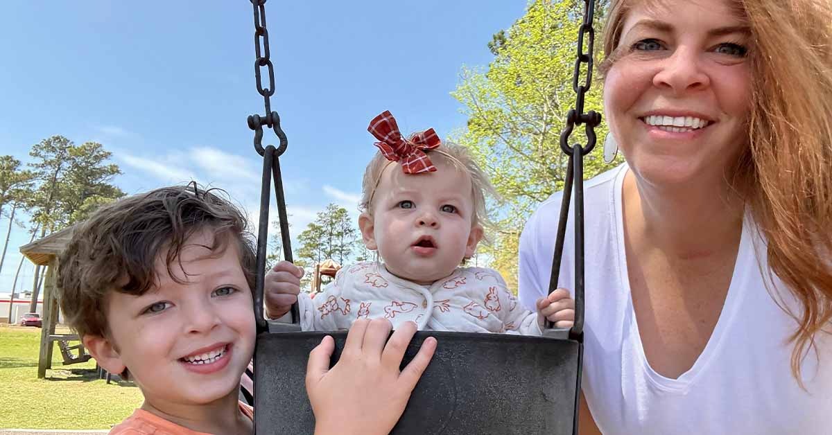 Lauren and two young children pose at a playground, with a baby in a swing and an older child gently pushing under a clear blue sky.