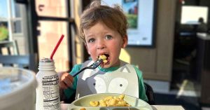 Lauren’s young child sits at a table eating a meal with a fork, wearing a bib, with a drink and plate of food.