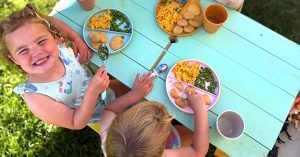 Children sit at a small outdoor table on grass, smiling and eating macaroni, peas and nuggets, with drinks beside them in bright sunlight.