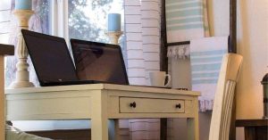 A home office desk by a window with a laptop, chair and natural light.