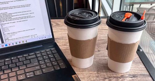 Laptop open beside two coffee cups on a wooden table by a window.