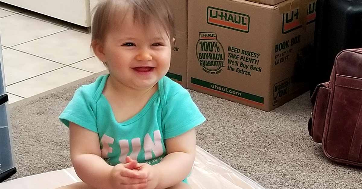 A smiling toddler sits in a room with moving boxes.