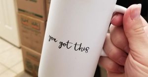 Hand holding a white mug with the text “you got this” in front of stacked moving boxes.