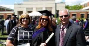 Lauren in a graduation cap and gown holding a diploma while posing outdoors with two smiling family members during a graduation ceremony.
