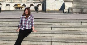 Lauren sitting on stone steps outside a government building, smiling while wearing a plaid shirt and black pants in bright daylight.