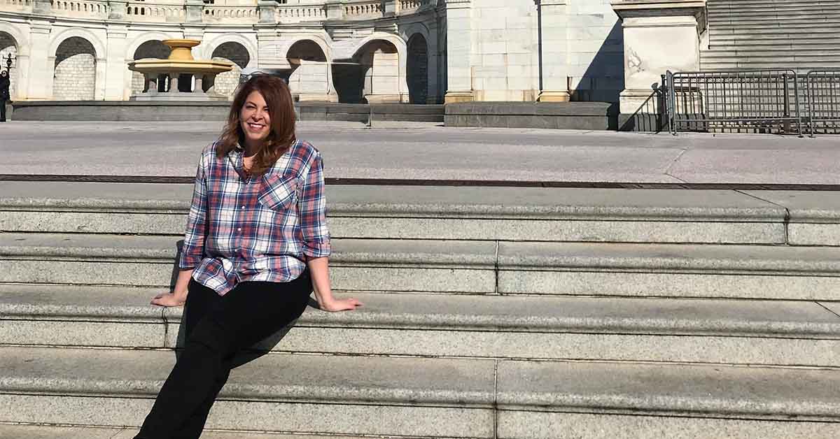 Lauren sitting on stone steps outside a government building, smiling while wearing a plaid shirt and black pants in bright daylight.