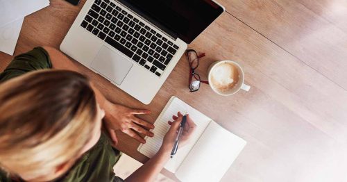 Person writing in a notebook beside a laptop, coffee cup, and glasses on a wooden desk, viewed from above.