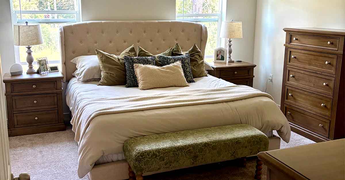 Neutral-toned bedroom with a tufted upholstered bed, layered decorative pillows, matching wooden nightstands, and natural light from two windows.
