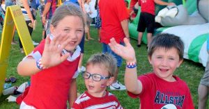 Three children waving and smiling at an outdoor community event with inflatable activities and crowds in the background.