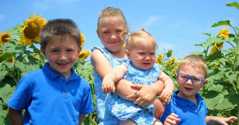 Lizann’s family smiling together in a sunflower field, with four children dressed in blue outfits under a bright summer sky.