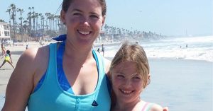 Lizann and her daughter smiling together on a sunny beach with ocean waves and palm trees in the background.