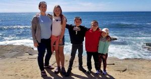 Lizann and her children posing together on a rocky ocean overlook with waves crashing behind them on a sunny day.