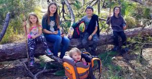 Lizann’s children sitting and climbing on a fallen tree in a wooded area, with a toddler seated in a child carrier backpack.