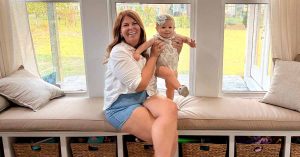 Lauren smiles while holding her baby as she sits on a cushioned window seat.