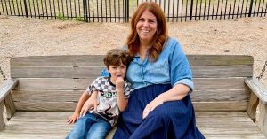Lauren smiles while sitting on a wooden bench with her son beside her.