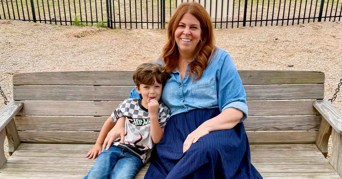 Lauren smiles while sitting on a wooden bench with her son beside her.