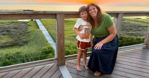 Lauren poses with her son on a wooden deck overlooking a grassy landscape at sunset.
