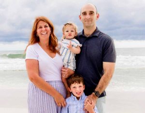 Lauren poses with her husband, son and a baby on a beach with ocean waves behind them.