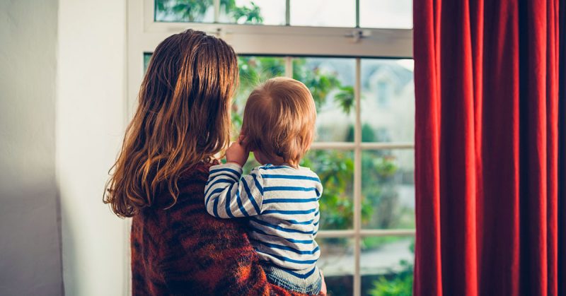 A woman holds a baby looking out a window.
