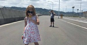 A girl in sunglasses and a patterned dress holds a flower on a wide road while a boy stands behind her with mountains in the distance.