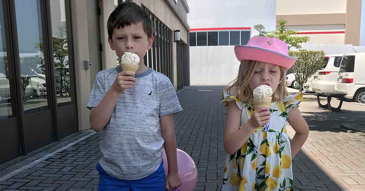 A boy and girl stand outdoors eating ice cream cones; the girl wears a pink cowboy hat and lemon-print dress.