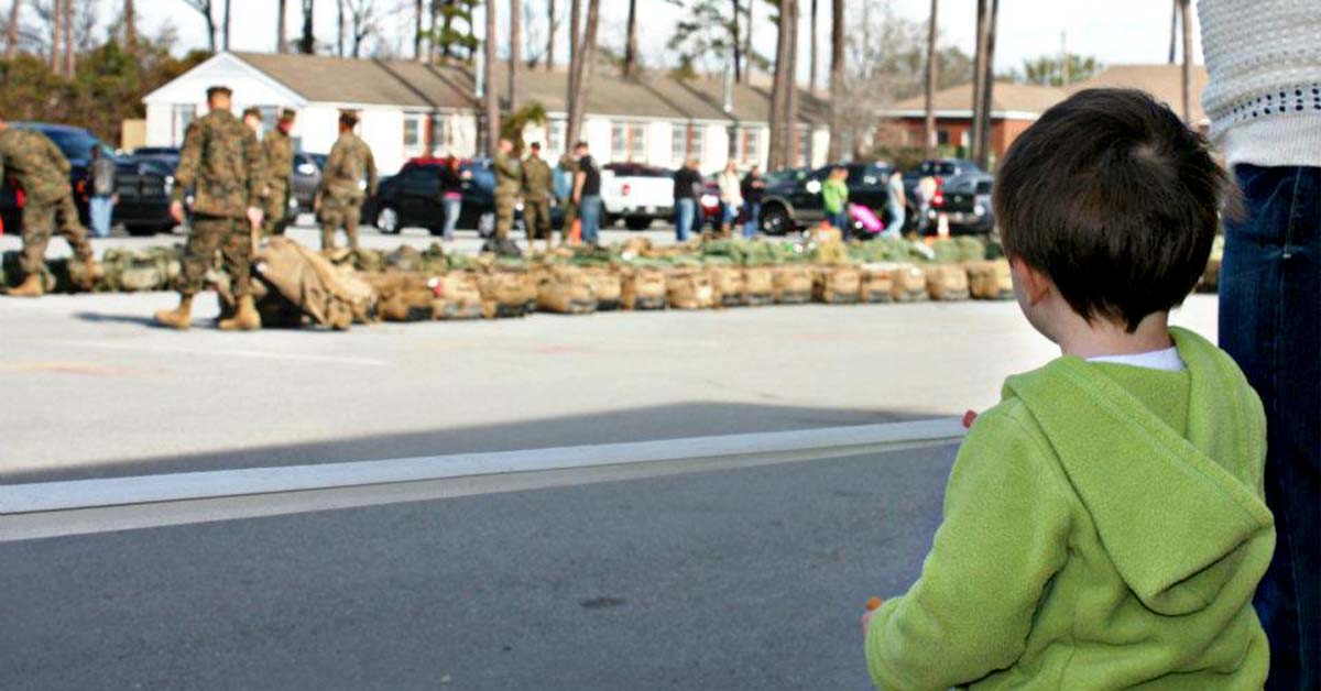 A young child watches uniformed service members and families gathered outdoors near rows of military gear.