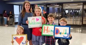 Lizann and four children stand at an airport holding handmade signs that read “Welcome,” “Home,” “Daddy,” and “We Love You!”