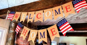 Red, white and blue “Welcome Home” banners hang with American flags in a rustic room.