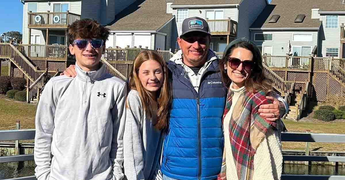 Kristi and her family smiling together outdoors in front of waterfront homes.
