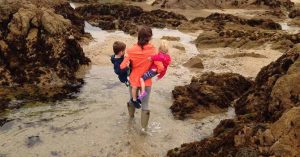 Kristi carrying her two young children through shallow water at a rocky beach. 