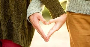 Two people form a heart shape with their hands while standing side by side outdoors.
