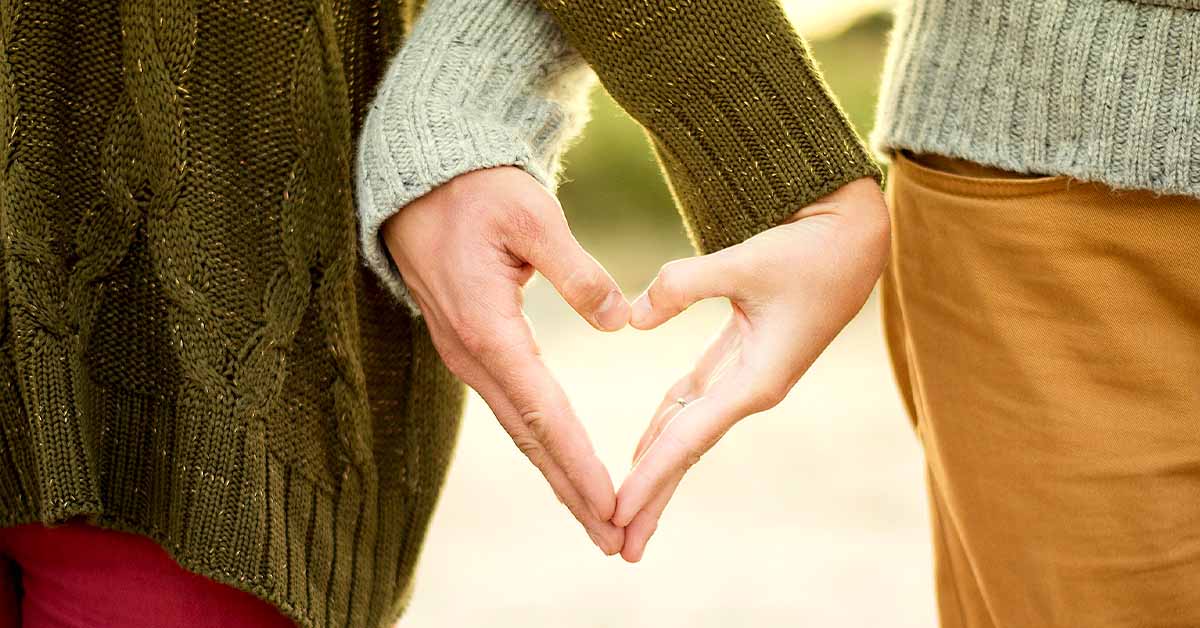 Two people form a heart shape with their hands while standing side by side outdoors.