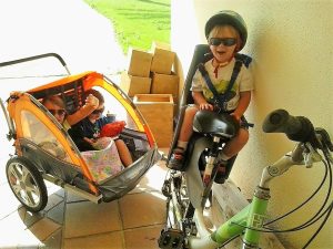 Lizann’s children riding in a bike trailer and child bike seat while wearing helmets and sunglasses during an outdoor family outing.