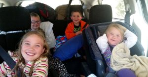 Four kids smile for the camera in a car that is loaded up with pillows and bags for a roadtrip.