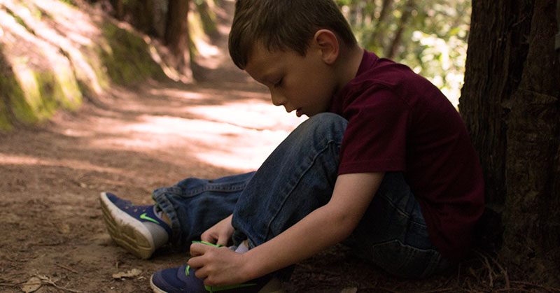 A boy sitting next to a tree tying his shoelaces